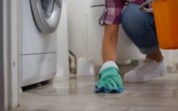 Crouching woman cleans up water from a leaking washer with a bucket and rag